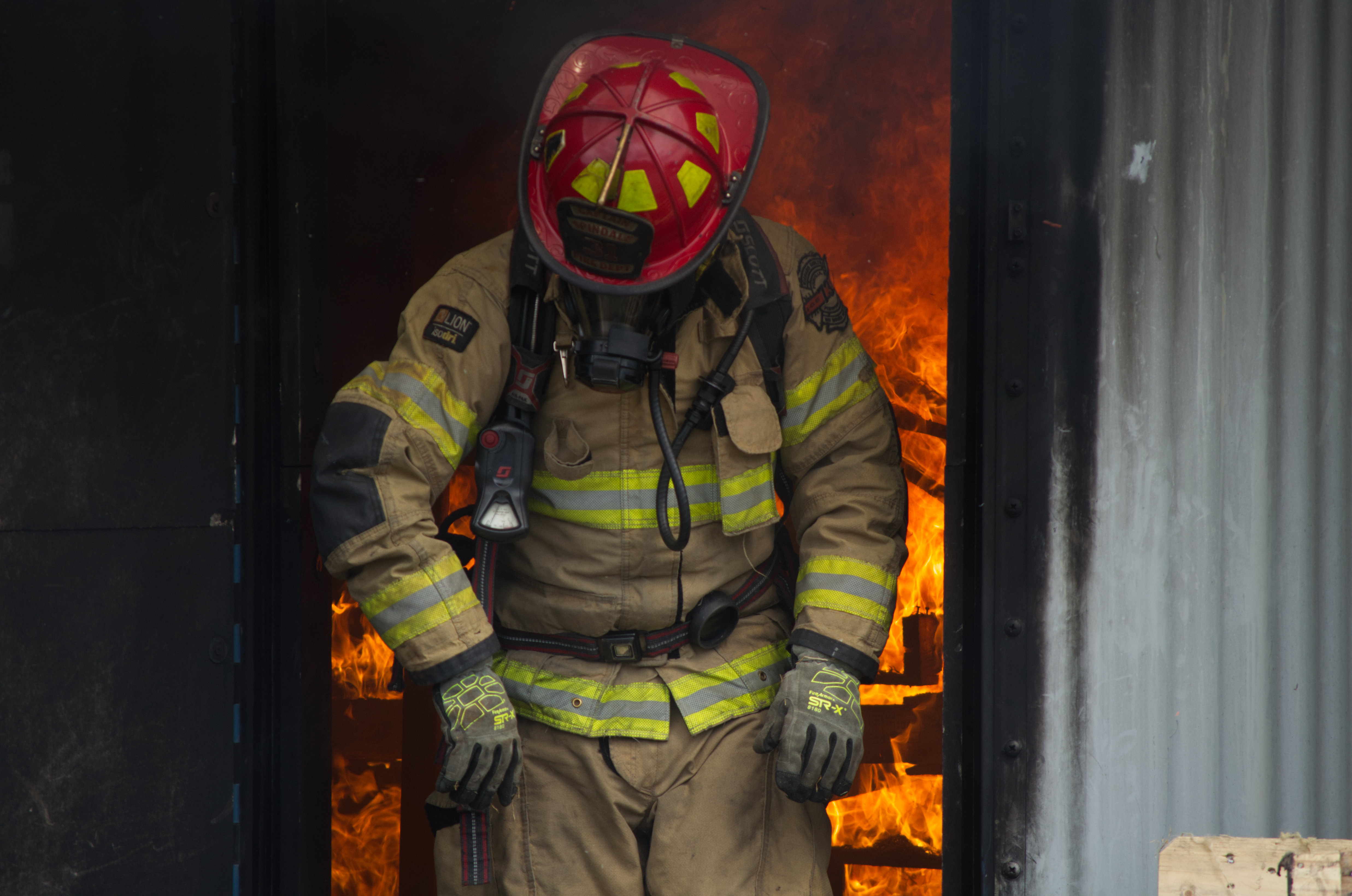 Fire fighter checking over his suit after walking out of burning building while flames are visible behind him