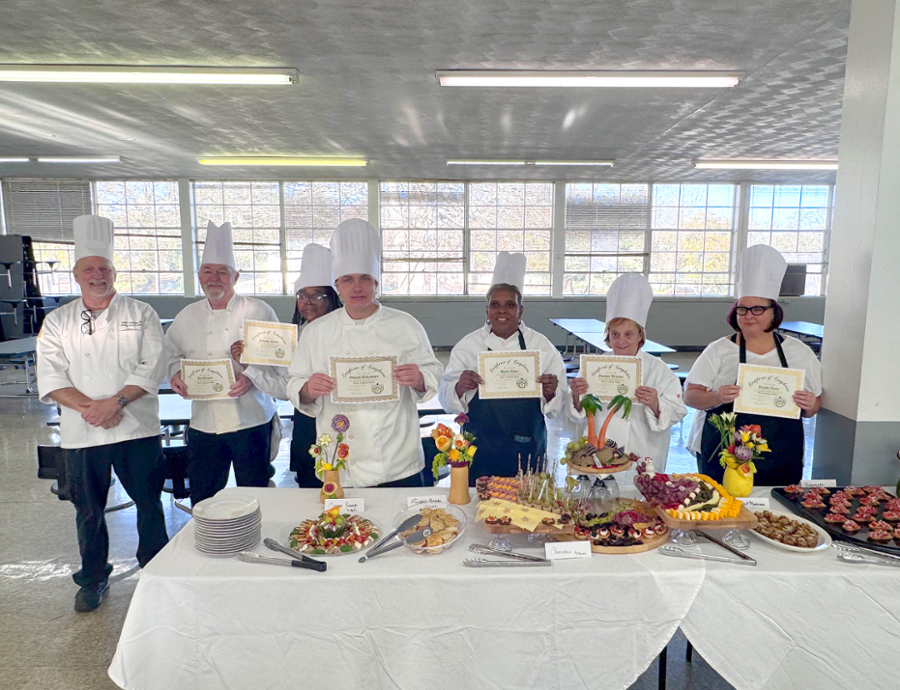 Group of chefs in white uniforms and tall hats holding certificates, standing behind a table displaying a variety of colorful dishes and desserts in a bright room with large windows.