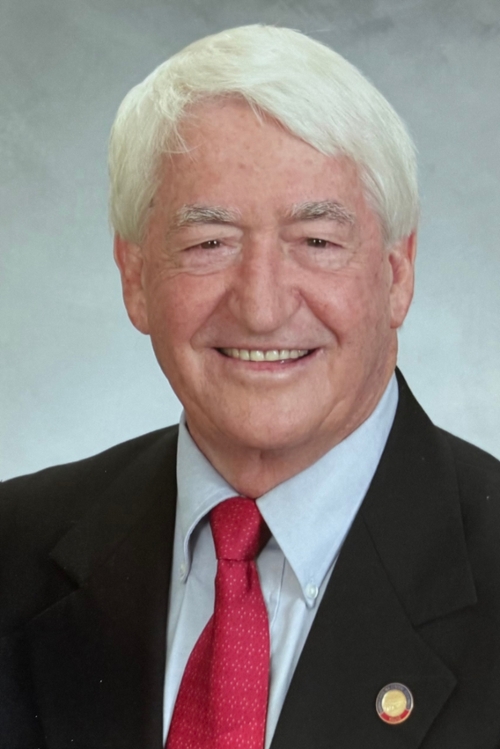 Headshot of Dr. Bob England. Smiling older man with white hair wearing a black suit, white shirt, red tie, and a lapel pin, posed against a gray background.