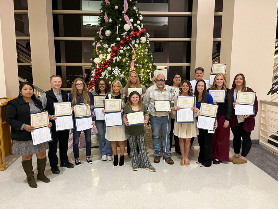 Group of people holding Student of the Semester awards certificates, standing in front of a decorated Christmas tree in a well-lit lobby.