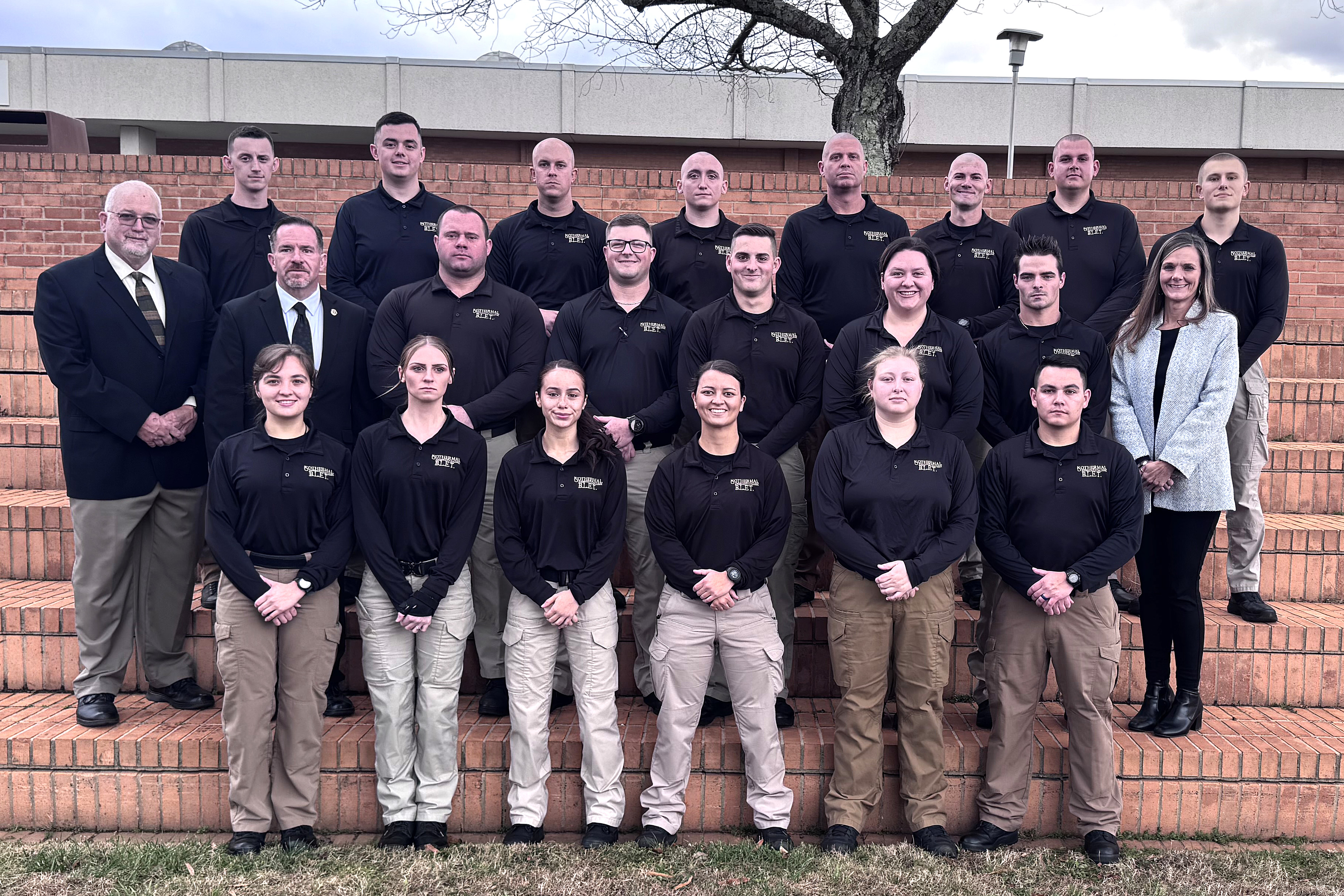 A group of individuals, including men and women, standing on brick steps outdoors for a formal group photo. Most wear black uniform shirts and khaki pants, while two men in the front row wear suits and ties, and one woman on the far right wears a light blue coat. A tree and building are visible in the background.